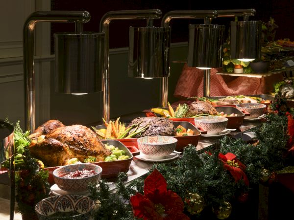 A festive buffet table with various dishes, including roasted meats and vegetables, decorated with greenery and flowers under warm lighting.