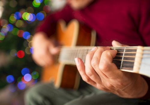 A person playing an acoustic guitar with a colorful, blurred background, possibly holiday lights, creating a warm, festive atmosphere.