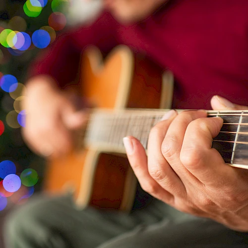 A person is playing an acoustic guitar with festive, colorful lights blurred in the background, creating a warm and cozy atmosphere.