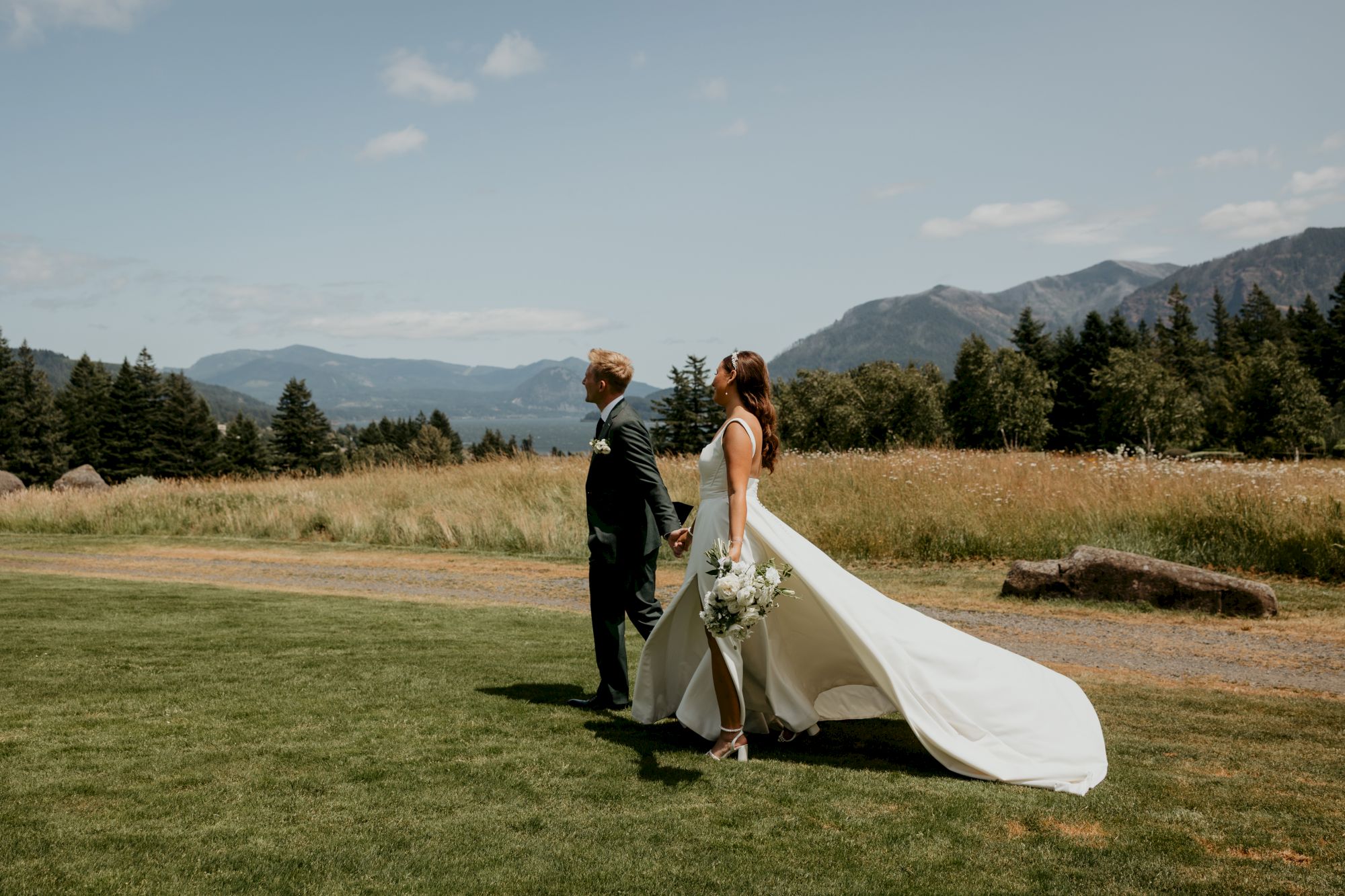 A couple in wedding attire walks in a scenic field with mountains and trees in the background, under a clear sky.