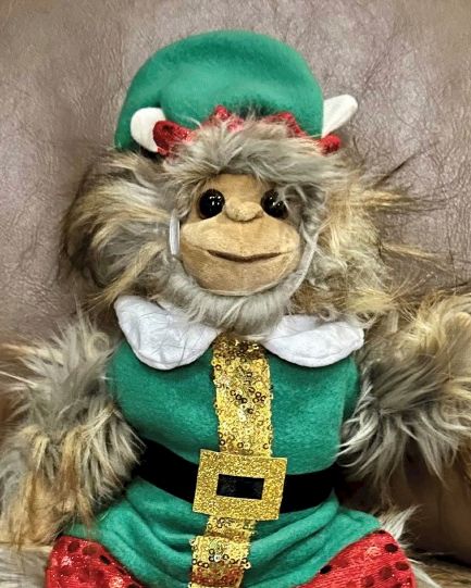 A furry toy dressed as an elf with a green hat, gold belt, and red pants, sitting on a brown surface.
