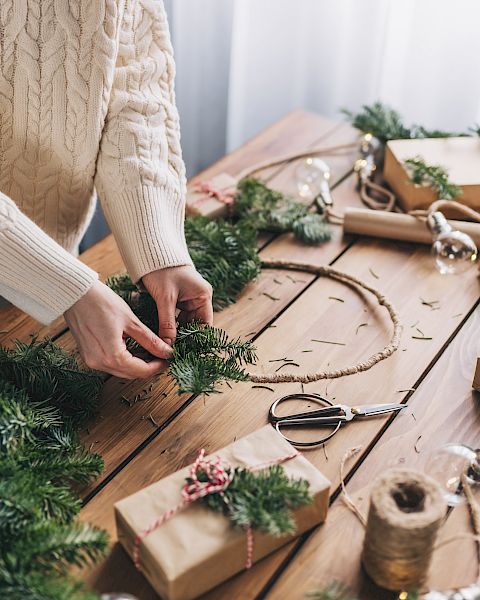 A person is crafting a holiday wreath at a table filled with wrapped gifts and greenery, creating a festive atmosphere.