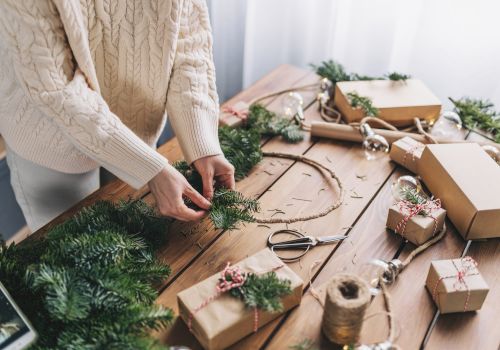 A person is crafting a holiday wreath at a table filled with wrapped gifts and greenery, creating a festive atmosphere.