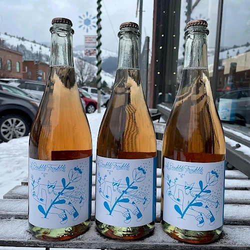 Three small glass bottles with light pink to amber liquid, labeled with blue botanical designs, on a snowy outdoor bench, reflective storefronts nearby.