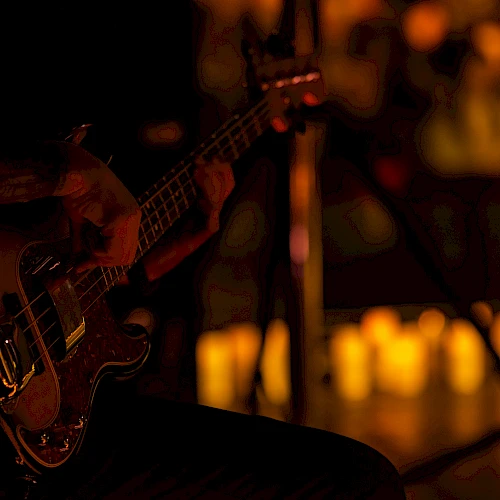 A dimly lit stage with a bass guitar resting on a musician&rsquo;s knee; warm, blurry background lights hint at an audience or venue.