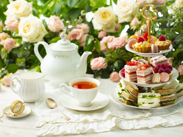 A delicate afternoon tea setup with a teapot, cup, and a three-tiered tray of assorted pastries against a bloom-filled backdrop.