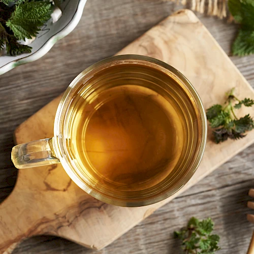 A cup of golden herbal tea sits on a wooden board with greens, a honey dipper, and fresh herbs around, cozy and rustic.