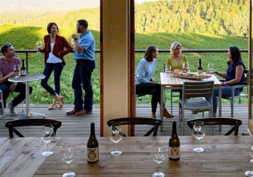People enjoy a twilight winery vibe on a terrace: groups chat, toast, and sip wine as the sun sets over green hills behind them.