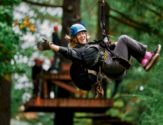 A person zip-lining through a forest, wearing a helmet and safety gear, smiling as they glide along a rope.
