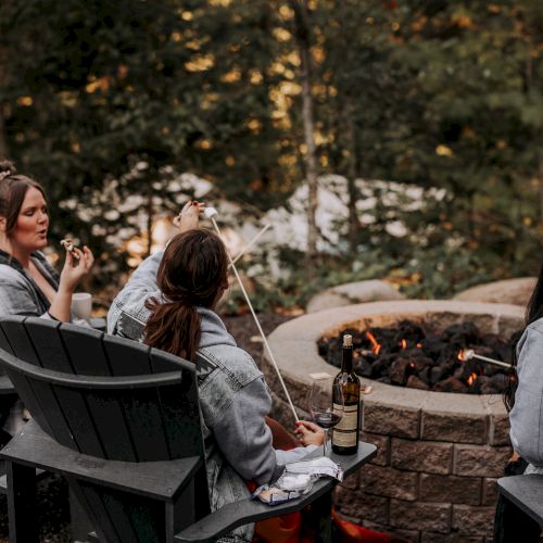 Three people sitting around a fire pit in a wooded area, roasting marshmallows, with one person holding a drink.