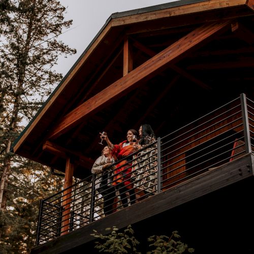 A group of people standing on a wooden balcony surrounded by trees, pointing at something in the distance, with a cabin structure above.