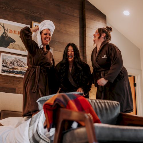Three people laughing and standing on a bed in a cozy room, all wearing brown robes; one has a towel on their head.