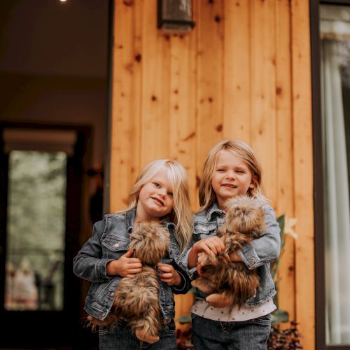 Two children in denim outfits are holding fluffy dogs in front of a wooden house, smiling cheerfully at the camera.