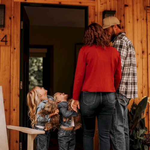 A family stands outside a wooden house; the children hold chickens and look up at the parents near an open door.