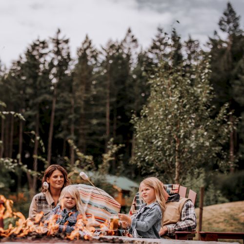 A group is sitting by a fire pit outdoors, surrounded by trees and relaxed atmosphere.