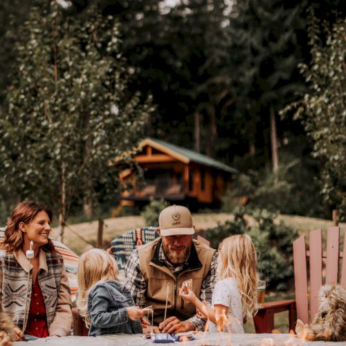 A family enjoys time together around a fire pit in a wooded area, with a cabin visible in the background, surrounded by trees.