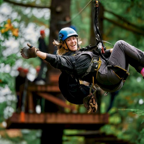 A person with a blue helmet is riding a zip line in a forest, smiling and waving, with others in the background on a platform.