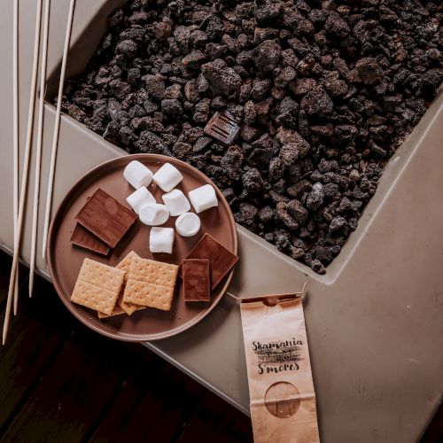A tray with graham crackers, chocolate, marshmallows, skewers, and a package labeled &ldquo;Signature Smores&rdquo; next to a coal pit.