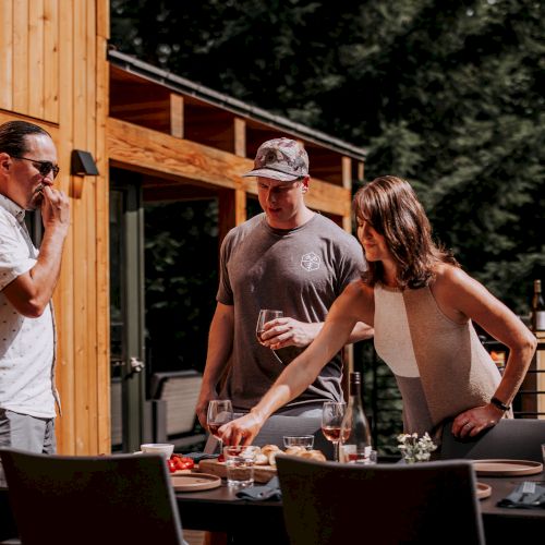 Three people are gathered outdoors by a wooden structure, enjoying drinks and snacks on a table, surrounded by trees in the background.