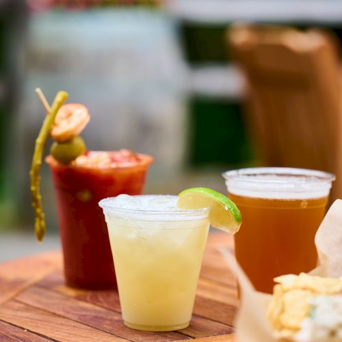 The image shows three drinks on a table, including a cocktail with olives and a lime, and a bowl of chips in the foreground.