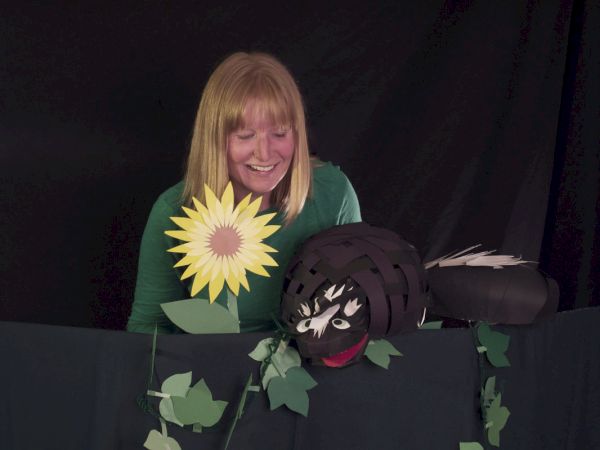 A person with a sunflower prop smiles behind a dark curtain, with a black puppet or mask-like creature peeking over the edge.