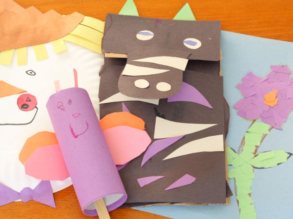 A colorful collage crafts scene: paper puppets and shapes on a wooden table, including a purple roll figure, a gray-face creature, and a blue card with a flower.