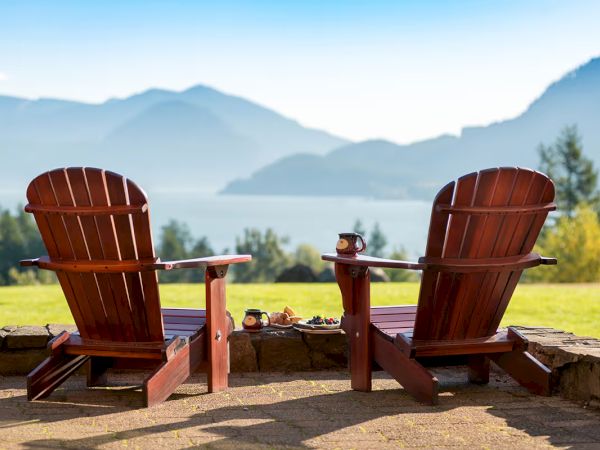 Two wooden chairs face a scenic view of mountains and a lake, with drinks placed on a stone ledge in front of them, under a clear sky.
