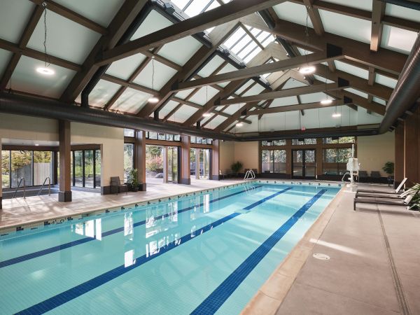 An indoor swimming pool with wooden beams, skylights, lounge chairs, and glass doors leading to an outdoor area.