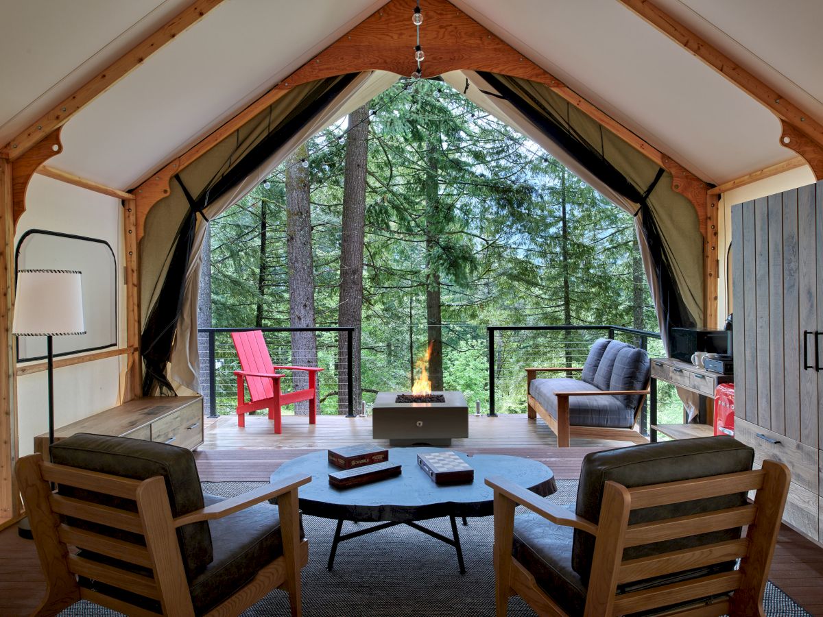 Cozy cabin living room with a circular seating arrangement, a central fire pit, wooden chairs, a terrace with forest views, and warm lighting.