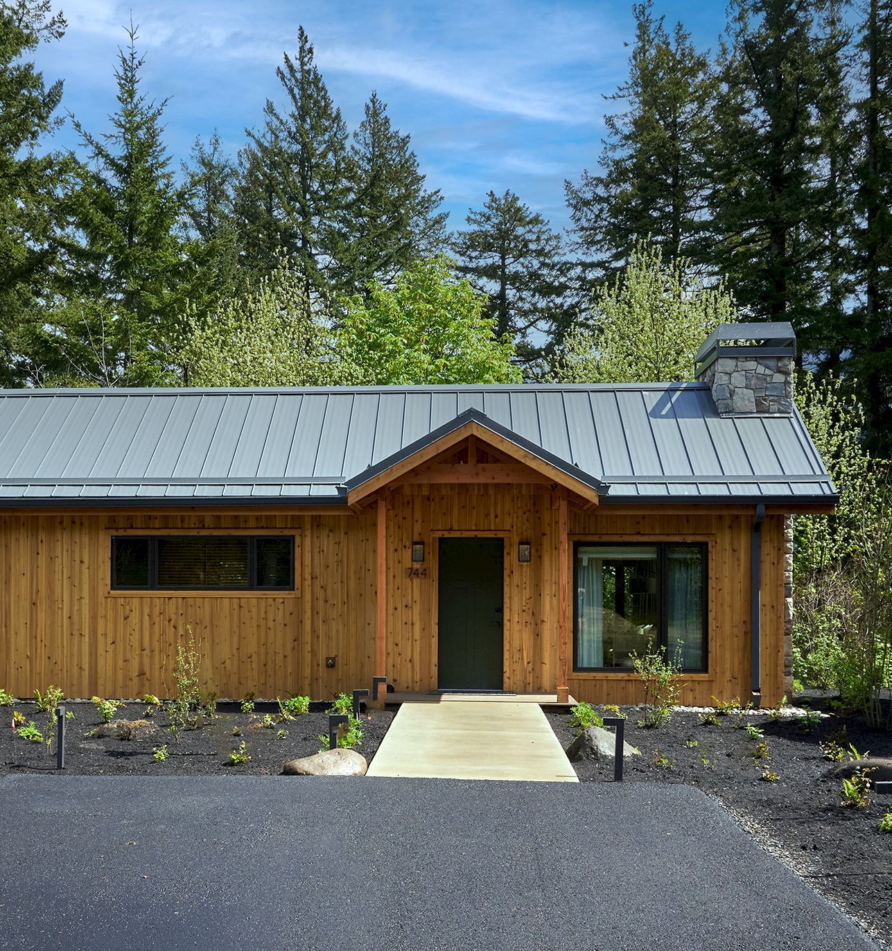 A modern wooden cabin with a dark metal roof, stone chimney, and a walkway in a lush green forest setting.