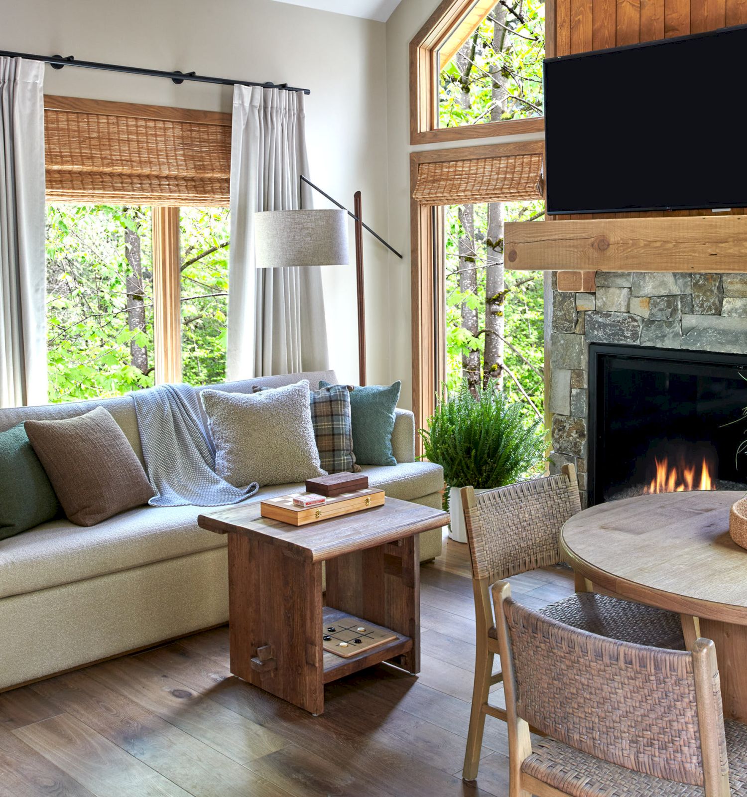 Cozy living room with a beige couch, stone fireplace with flames, large TV, and curtains framing green forest views.