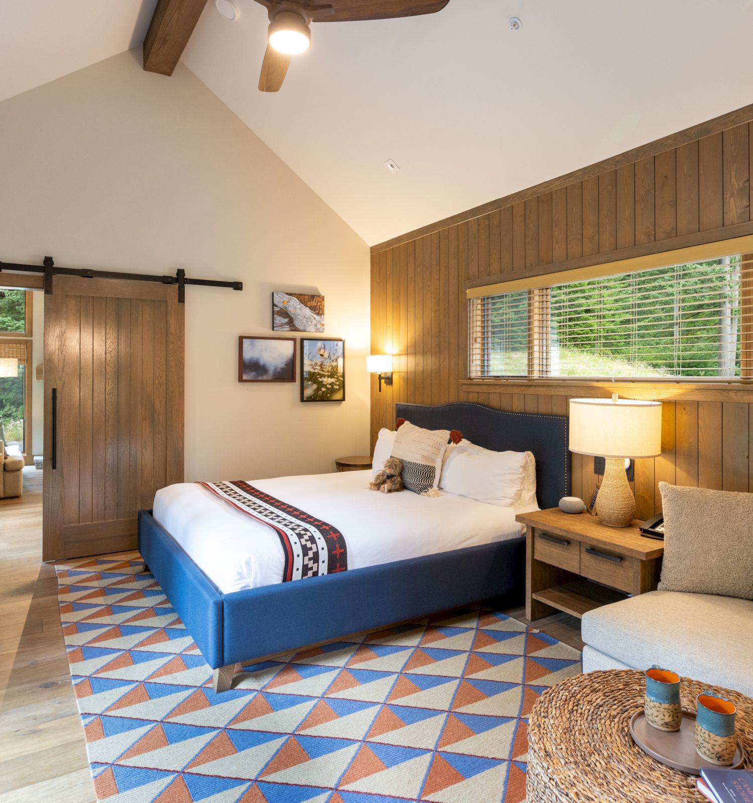 A cozy bedroom with a blue bed, wood paneling, large window blinds, a ceiling fan light, and a small seating area.