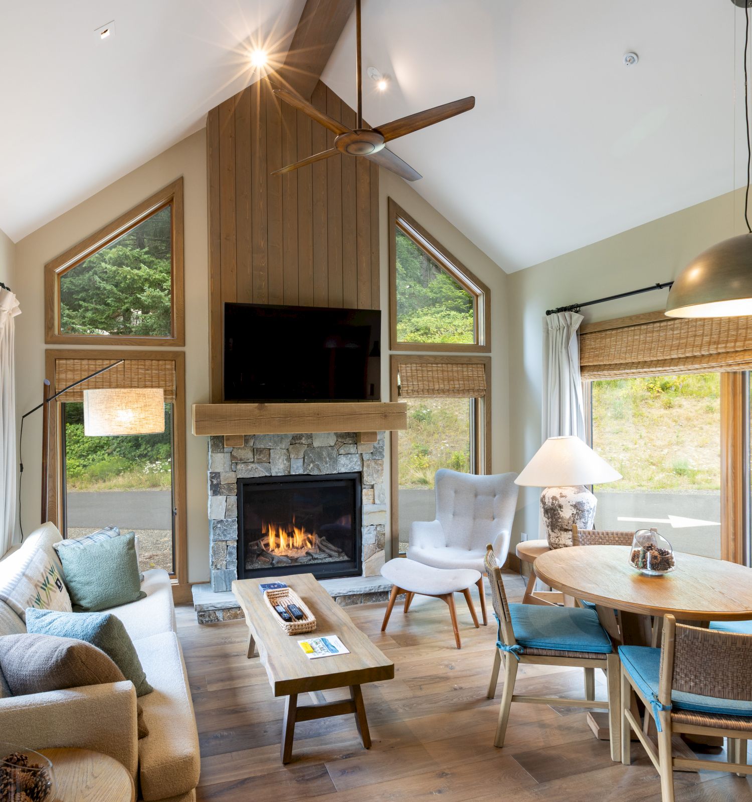Cozy living room with a stone fireplace, TV, large windows, ceiling fan, and comfy seating.