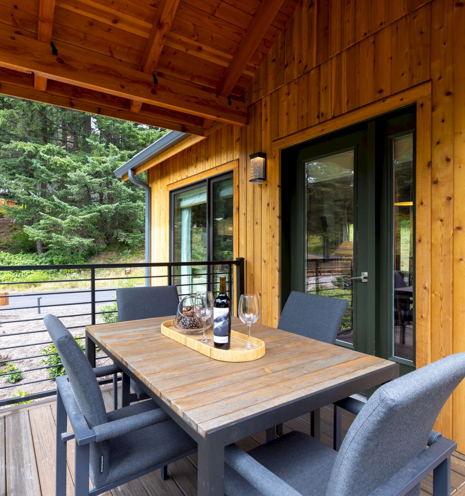A wooden balcony beside a cabin, with a table set with wine and glasses and gray chairs overlooking green forest views.