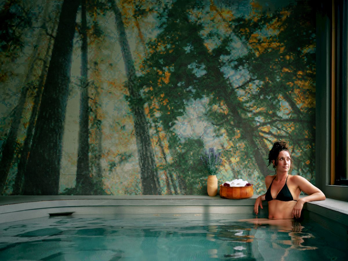 A woman in a spa pool lounges beside a mosaic-tiled wall with a vase and towels nearby, creating a calm wellness scene.