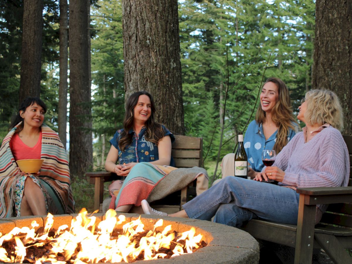 Friends sit on a park bench around a crackling outdoor fire in a green forest, laughing and sipping wine.