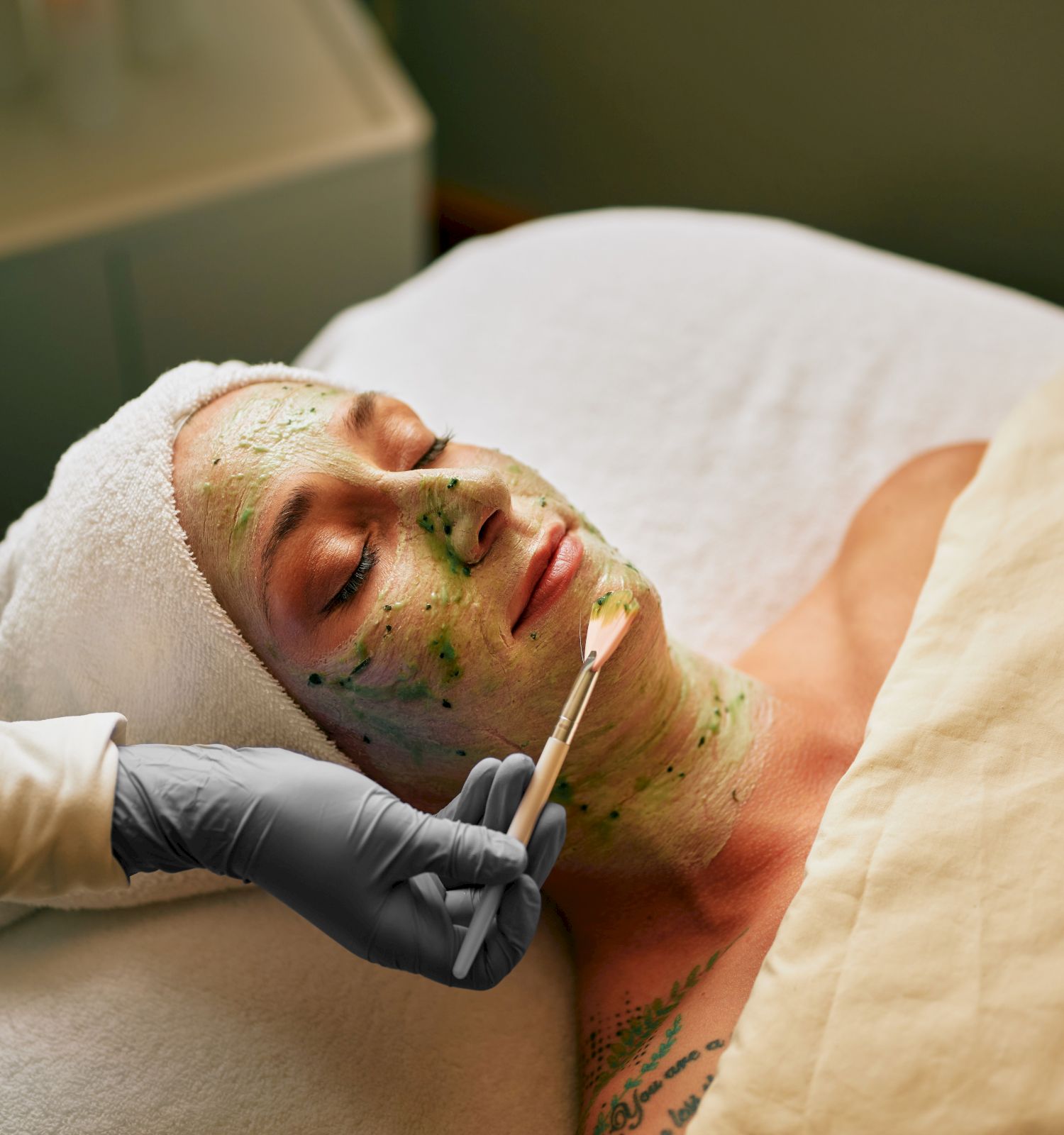 A facial treatment scene: a person lies on a bed with a green mask while a gloved clinician applies it with a tool.