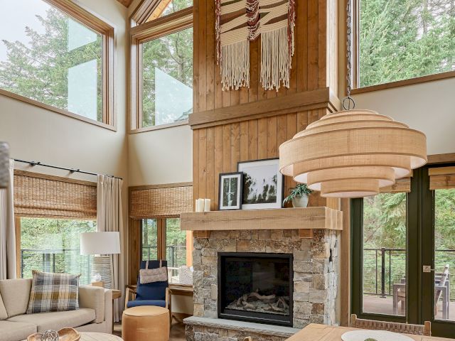 Cozy wooden living room with a stone fireplace, large windows to the forest, woven wall hangings, and warm hanging lamps.