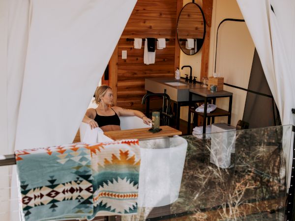A woman lounges in a modern wooden bathroom/vanity area with a sink, towels, and a large oval mirror, looking outside.