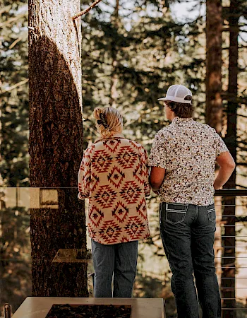 A couple enjoys a cozy cabin view in the forest, standing on a deck by a grill and benches at sunset.