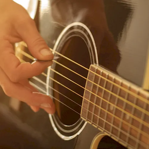 A person&rsquo;s hand plucks the strings of an acoustic guitar in a warm, close-up shot.