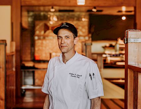 A chef in a white coat and cap stands in a rustic kitchen, with warm lights and a brick oven in the background.