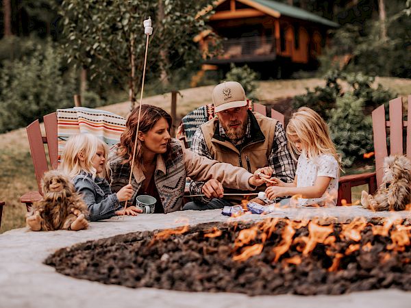 A family camping by a fire, roasting snacks at a picnic table with kids and dogs in a forest backyard.