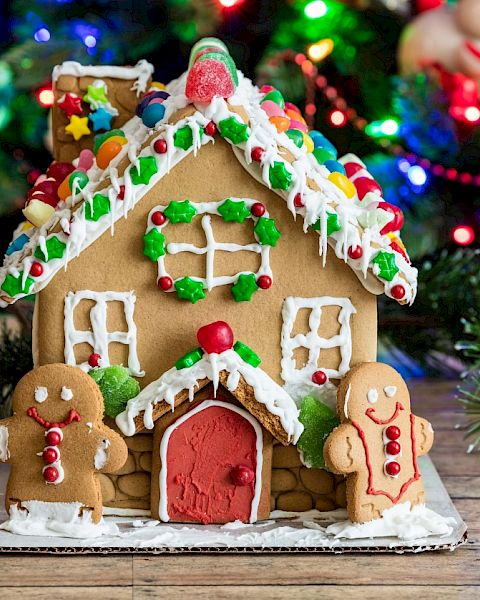 A decorated gingerbread house with gumdrop shingles and icing details sits on a holiday table, surrounded by colorful Christmas lights.