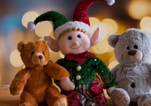 A smiling elf in a green sequined suit sits with three plush teddy bears&mdash;brown, gray, and cozy at a holiday table.
