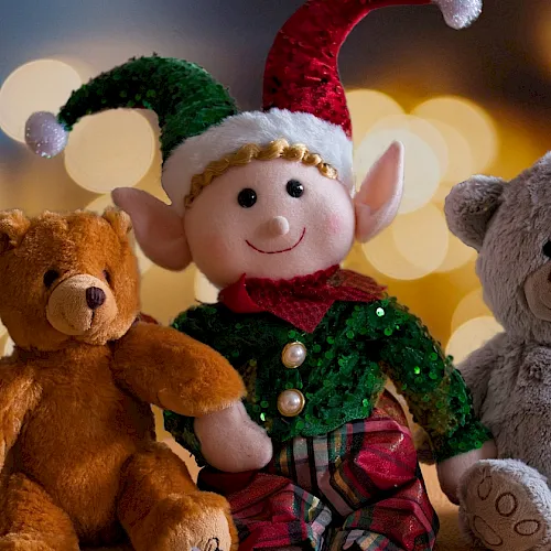 A smiling elf in a green sequined suit sits with three plush teddy bears&mdash;brown, gray, and cozy at a holiday table.