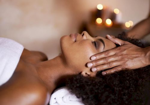 A relaxing couple&rsquo;s massage: a therapist gently holds a woman&rsquo;s head and face with warm, cozy lights in the background.