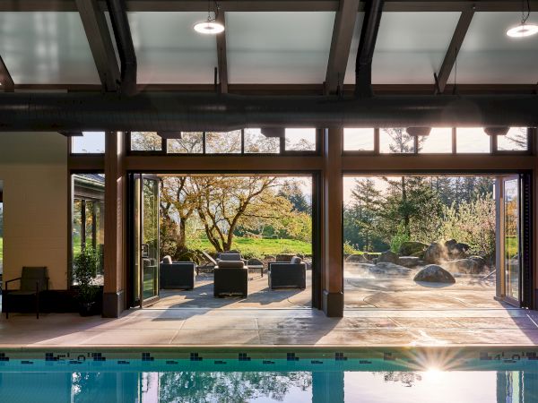 The image shows an indoor pool area with open glass doors leading to an outdoor seating area and garden, bathed in sunlight.