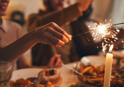 A hand holds a lit sparkler over a festive dinner table with a candle and people blurred in the background.
