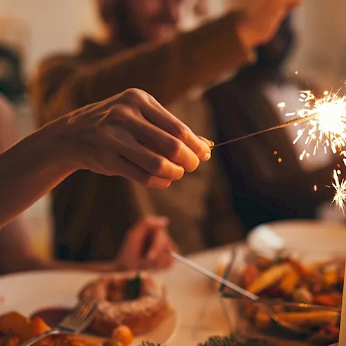 A hand holds a lit sparkler over a festive dinner table with a candle and people blurred in the background.
